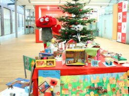 Lotties Welt: 30 Gewinner bei der Aktion „Wunschbaum“ Das Pilz-Maskottchen der Lotterie Los-Sparen steht vor einem Weihnachtsbaum. Vor ihm ist ein Tisch voller Basteleien von Kindern.