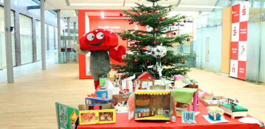Lotties Welt: 30 Gewinner bei der Aktion „Wunschbaum“ Das Pilz-Maskottchen der Lotterie Los-Sparen steht vor einem Weihnachtsbaum. Vor ihm ist ein Tisch voller Basteleien von Kindern.