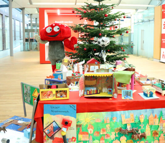 Lotties Welt: 30 Gewinner bei der Aktion „Wunschbaum“ Das Pilz-Maskottchen der Lotterie Los-Sparen steht vor einem Weihnachtsbaum. Vor ihm ist ein Tisch voller Basteleien von Kindern.