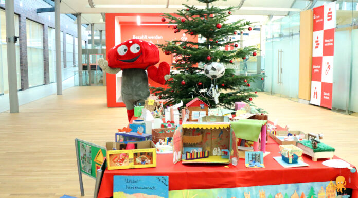Lotties Welt: 30 Gewinner bei der Aktion „Wunschbaum“ Das Pilz-Maskottchen der Lotterie Los-Sparen steht vor einem Weihnachtsbaum. Vor ihm ist ein Tisch voller Basteleien von Kindern.