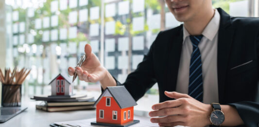 Warum Bausparverträge ein Comeback erleben Ein Bankkaufmann sitzt am Schreibtisch mit einem Vertrag, auf dem ein kleines rotes Haus steht.