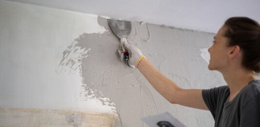 Muskelhypothek – beim Hausbau anpacken und Geld sparen Junge Frau, die Hausreparaturen durch Auftragen eines Pflasters mit einem Tuch durchführt.