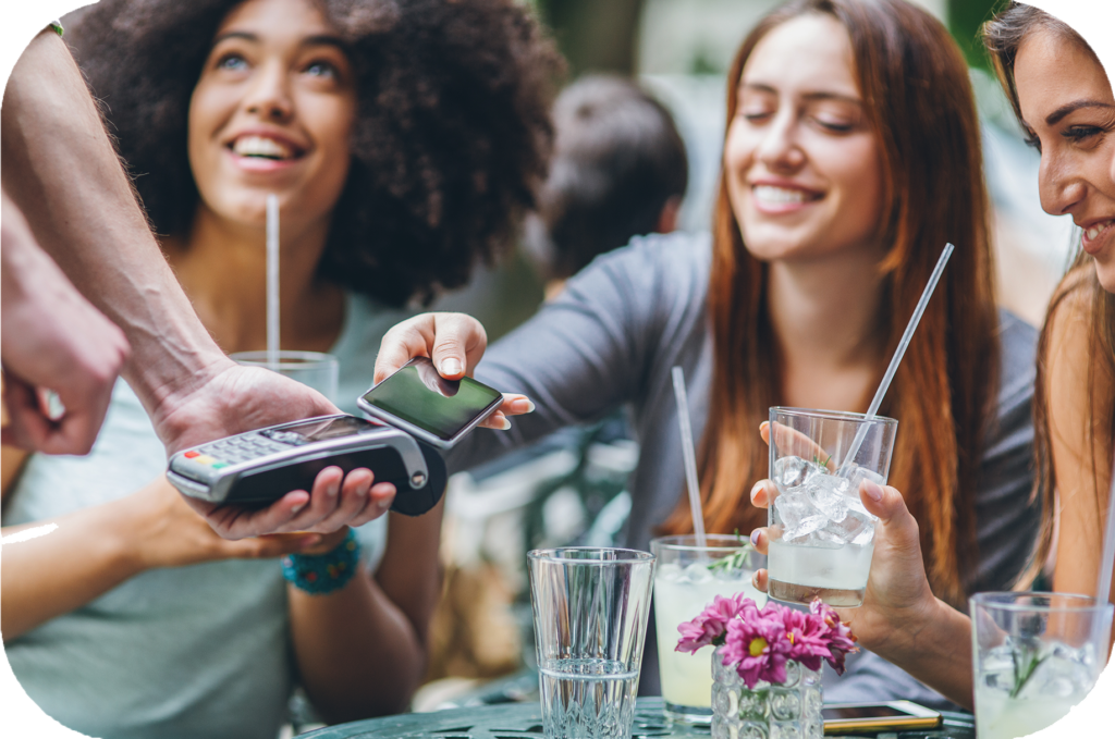 Drei junge Frauen im Café, eine bezahlt mit ihrem Smartphone.
