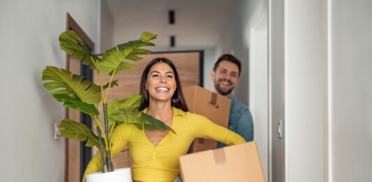 Bezahlbarer Wohnraum – eine Übersicht Junges Ehepaare zieht in neues Zuhause um mit Kartons und Pflanze.