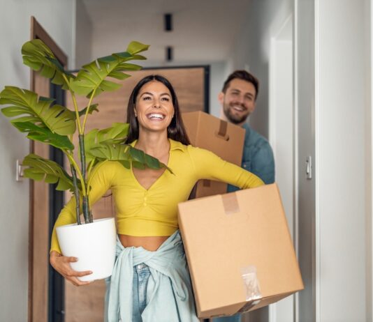 Bezahlbarer Wohnraum – eine Übersicht Junges Ehepaare zieht in neues Zuhause um mit Kartons und Pflanze.