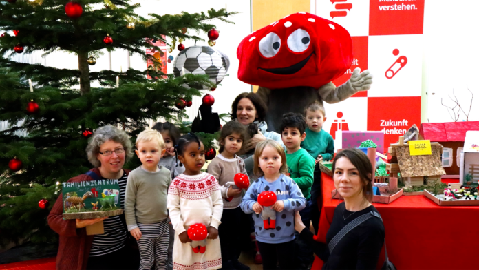 Fliegenpilz Maskottchen steht mit Kindern neben einem Weihnachtsbaum.