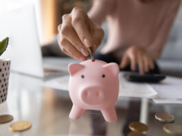 Notgroschen: Wie viel Geld sollte ich beiseitelegen? Nahaufnahme von junger Frau, die Münze im Sparschwein einsetzt.