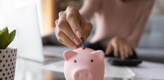 Notgroschen: Wie viel Geld sollte ich beiseitelegen? Nahaufnahme von junger Frau, die Münze im Sparschwein einsetzt.