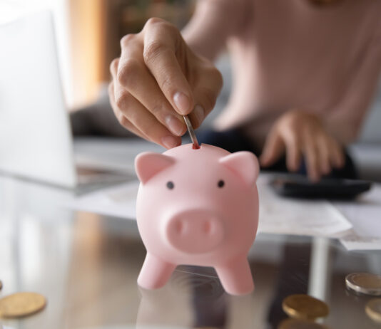 Notgroschen: Wie viel Geld sollte ich beiseitelegen? Nahaufnahme von junger Frau, die Münze im Sparschwein einsetzt.