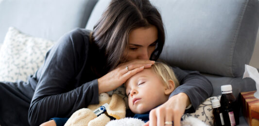 Kinderkrankengeld: Das sind Ihre Ansprüche Kleiner blonder Junge schläft im Arm seiner Mutter auf dem Sofa, daneben Medikamente