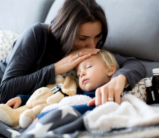 Kinderkrankengeld: Das sind Ihre Ansprüche Kleiner blonder Junge schläft im Arm seiner Mutter auf dem Sofa, daneben Medikamente