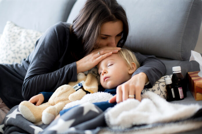 Kleiner blonder Junge schläft im Arm seiner Mutter auf dem Sofa, daneben Medikamente
