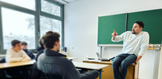 Tatort soziale Netzwerke: Verschwörungsmythen und Fake News im Internet Ein Mann sitzt vor einer Schulklasse auf einem Tisch vor einer Tafel.