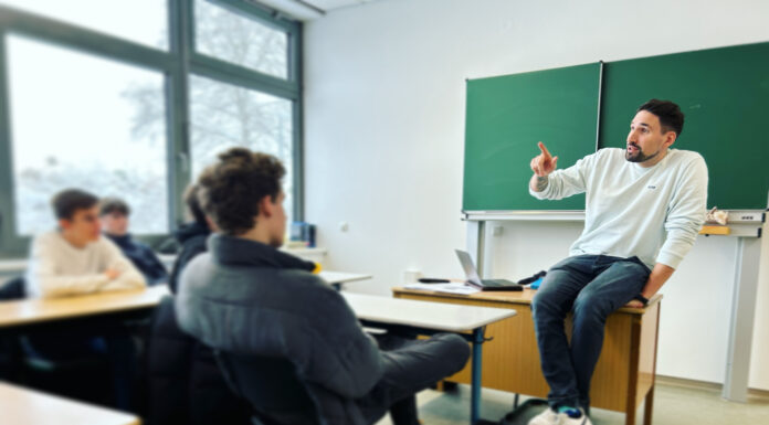 Tatort soziale Netzwerke: Verschwörungsmythen und Fake News im Internet Ein Mann sitzt vor einer Schulklasse auf einem Tisch vor einer Tafel.