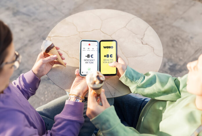 Zwei Menschen sitzen nebeneinander und haben Eistüten in der Hand, sie halten ihre Smartphones nebeneinander und bezahlen mit dem neuen Bezahlsystem wero