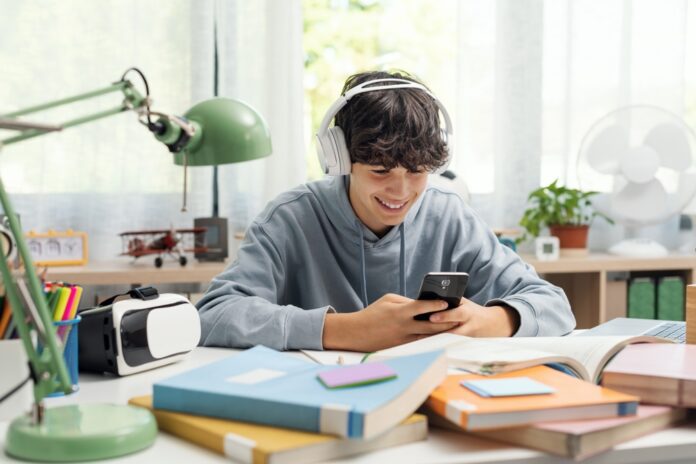 Teenager sitzt zuhause am Schreibtisch und hat Kopfhörer auf und ein Smartphone in der Hand