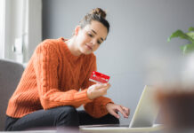 Sichere Online-Zahlung mit der neuen Sparkassen-Card Frau im orangenen Pullover sitzt auf den Sofa und hält eine Sparkassen Card in der Hand, vor ihr steht ein Laptop