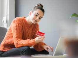 Sichere Online-Zahlung mit der neuen Sparkassen-Card Frau im orangenen Pullover sitzt auf den Sofa und hält eine Sparkassen Card in der Hand, vor ihr steht ein Laptop