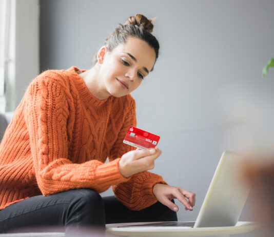 Sichere Online-Zahlung mit der neuen Sparkassen-Card Frau im orangenen Pullover sitzt auf den Sofa und hält eine Sparkassen Card in der Hand, vor ihr steht ein Laptop