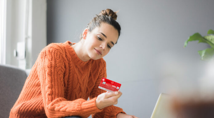 Sichere Online-Zahlung mit der neuen Sparkassen-Card Frau im orangenen Pullover sitzt auf den Sofa und hält eine Sparkassen Card in der Hand, vor ihr steht ein Laptop