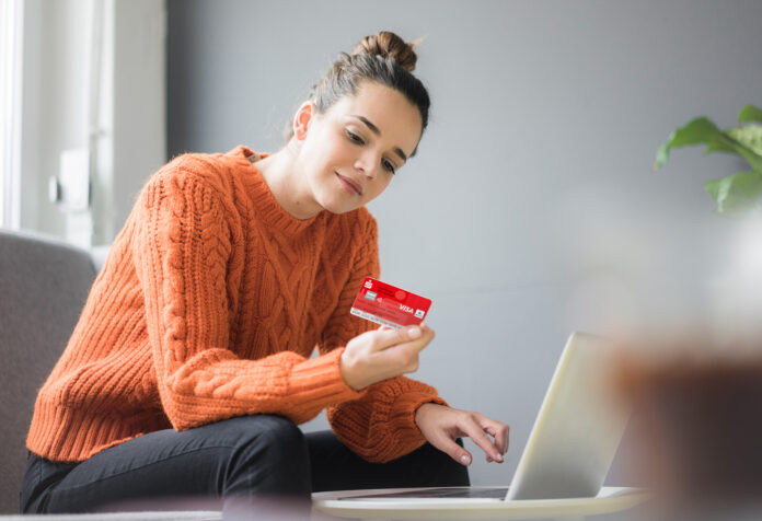 Frau im orangenen Pullover sitzt auf den Sofa und hält eine Sparkassen Card in der Hand, vor ihr steht ein Laptop