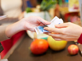 Bargeldservice in Supermärkten & Co. Eine Hand reicht an einer Supermarkt Kasse Euroscheine an eine andere Hand weiter.