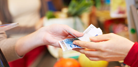 Bargeldservice in Supermärkten & Co. Eine Hand reicht an einer Supermarkt Kasse Euroscheine an eine andere Hand weiter.