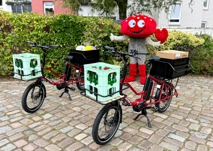 Zwei rote Lastenräder mit jeweils einer Kiste mit Wasserflaschen vorne dran. Dahinter steht ein Maskottchen in Form eines roten Fliegenpilzes.