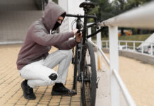 Fahrraddiebstähle: Schützen Sie sich finanziell mit einer Fahrradversicherung Ein Dieb in einer Balaclava und mit einer Kapuze bricht das Schloss an einem Fahrrad auf der Straße auf.