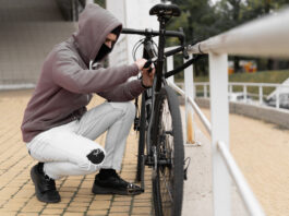 Fahrraddiebstähle: Schützen Sie sich finanziell mit einer Fahrradversicherung Ein Dieb in einer Balaclava und mit einer Kapuze bricht das Schloss an einem Fahrrad auf der Straße auf.