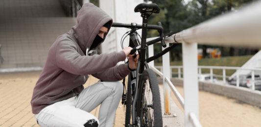 Fahrraddiebstähle: Schützen Sie sich finanziell mit einer Fahrradversicherung Ein Dieb in einer Balaclava und mit einer Kapuze bricht das Schloss an einem Fahrrad auf der Straße auf.