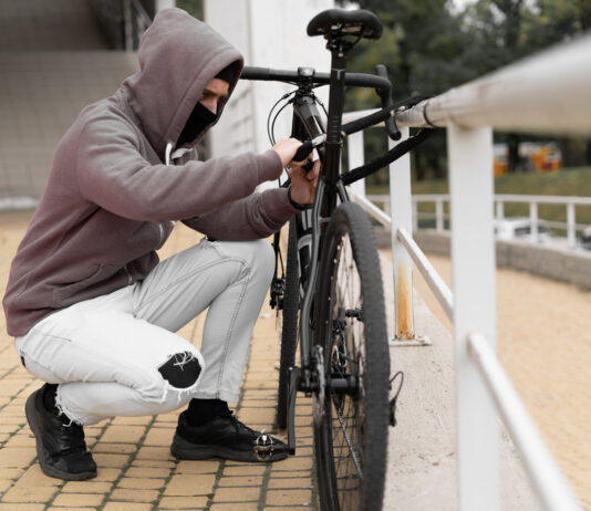Fahrraddiebstähle: Schützen Sie sich finanziell mit einer Fahrradversicherung Ein Dieb in einer Balaclava und mit einer Kapuze bricht das Schloss an einem Fahrrad auf der Straße auf.