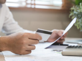 Auslagen und Spesen – 8 Tipps für mehr Effizienz in Unternehmen Ein Mann sitzt am Schreibtisch und hält eine Kreditkarte und Rechnungen in der Hand.