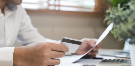 Auslagen und Spesen – 8 Tipps für mehr Effizienz in Unternehmen Ein Mann sitzt am Schreibtisch und hält eine Kreditkarte und Rechnungen in der Hand.