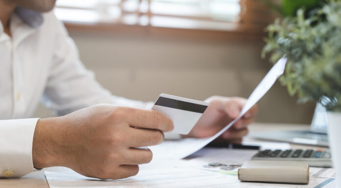 Auslagen und Spesen – 8 Tipps für mehr Effizienz in Unternehmen Ein Mann sitzt am Schreibtisch und hält eine Kreditkarte und Rechnungen in der Hand.