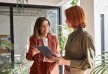 Neue Wirtschafts-Identifikationsnummer für Unternehmen kommt ab Herbst Zwei glückliche Geschäftsfrauen im mittleren und jungen Alter, die in kreativen, grünen Büroräumen stehen und mit digitalen Tablets bei der Arbeit Gespräche führen.