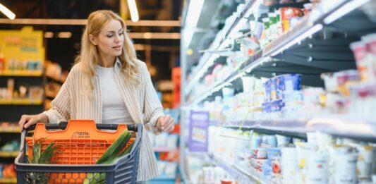 Inflationsrate: Was wurde wie viel teurer seit 2019? Junge blonde Frau, die Produkte im Kühlregal eines Lebensmittelgeschäftes anschaut.