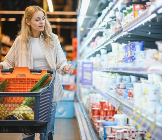 Inflationsrate: Was wurde wie viel teurer seit 2019? Junge blonde Frau, die Produkte im Kühlregal eines Lebensmittelgeschäftes anschaut.