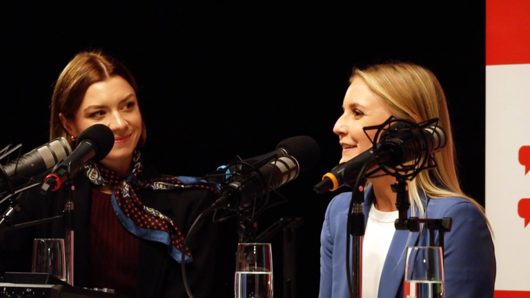 Eine brünette und eine blonde Frau sitzen hinter Mikrofonen und nehmen einen Podcast auf.