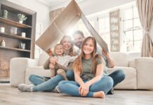 Miete und Eigenheim 2025: Das ändert sich rund um Ihre Immobilie Eine Mutter und ein Vater sitzen mit ihrem kleinen Sohn und der Teenager Tochter lächelnd in ihrem Wohnzimmer auf dem Fußboden und halten ein Pappdach über sich.