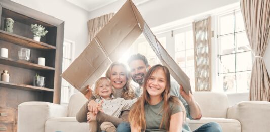 Miete und Eigenheim 2025: Das ändert sich rund um Ihre Immobilie Eine Mutter und ein Vater sitzen mit ihrem kleinen Sohn und der Teenager Tochter lächelnd in ihrem Wohnzimmer auf dem Fußboden und halten ein Pappdach über sich.