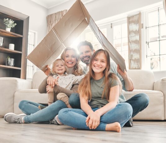 Miete und Eigenheim 2025: Das ändert sich rund um Ihre Immobilie Eine Mutter und ein Vater sitzen mit ihrem kleinen Sohn und der Teenager Tochter lächelnd in ihrem Wohnzimmer auf dem Fußboden und halten ein Pappdach über sich.
