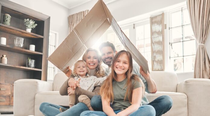 Miete und Eigenheim 2025: Das ändert sich rund um Ihre Immobilie Eine Mutter und ein Vater sitzen mit ihrem kleinen Sohn und der Teenager Tochter lächelnd in ihrem Wohnzimmer auf dem Fußboden und halten ein Pappdach über sich.