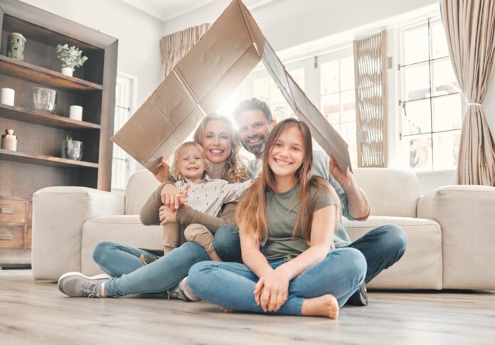 Eine Mutter und ein Vater sitzen mit ihrem kleinen Sohn und der Teenager Tochter lächelnd in ihrem Wohnzimmer auf dem Fußboden und halten ein Pappdach über sich.