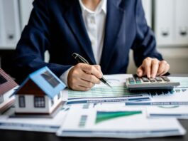 Neue Grundsteuer: Was Sie jetzt wissen und tun müssen Frau sitzt am Schreibtisch im Büro, tippt etwas in einen Taschenrechner ein. Auf dem Tisch liegen Akten und es stehen zwei Miniaturhäuser darauf.