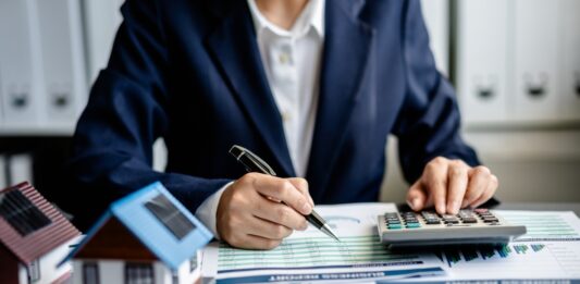 Neue Grundsteuer: Was Sie jetzt wissen und tun müssen Frau sitzt am Schreibtisch im Büro, tippt etwas in einen Taschenrechner ein. Auf dem Tisch liegen Akten und es stehen zwei Miniaturhäuser darauf.
