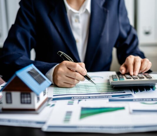 Neue Grundsteuer: Was Sie jetzt wissen und tun müssen Frau sitzt am Schreibtisch im Büro, tippt etwas in einen Taschenrechner ein. Auf dem Tisch liegen Akten und es stehen zwei Miniaturhäuser darauf.