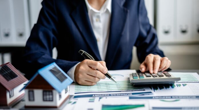 Neue Grundsteuer: Was Sie jetzt wissen und tun müssen Frau sitzt am Schreibtisch im Büro, tippt etwas in einen Taschenrechner ein. Auf dem Tisch liegen Akten und es stehen zwei Miniaturhäuser darauf.