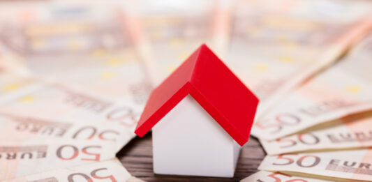 Günstiger ins Eigenheim: 10 Sparfaktoren Spielzeughaus mit rotem Dach steht im Zentrum von gefächerten 50-Euro-Scheinen.