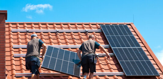 Photovoltaik: Wann sich Strom vom Dach lohnt Zwei Arbeiter installieren eine Solarzelle auf einem roten Ziegeldach.